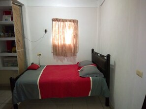 A single bed with a red and grey bedspread, a wooden headboard, and a window with sheer curtains.