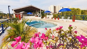 Una piscina con zona de estar cubierta, sillones reclinables blancos y sombrillas azules.