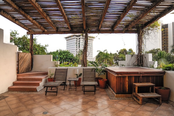 A covered outdoor area with a hot tub, wooden chairs, and potted plants.