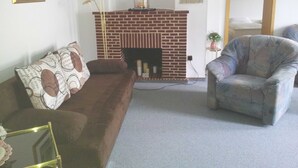 Una sala de estar con chimenea de ladrillo, un sofá marrón, un sillón estampado y una lámpara de pie.