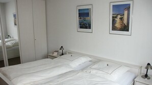 Una cama bien hecha con ropa blanca, una mesita de noche con lámpara y dos cuadros en la pared.
