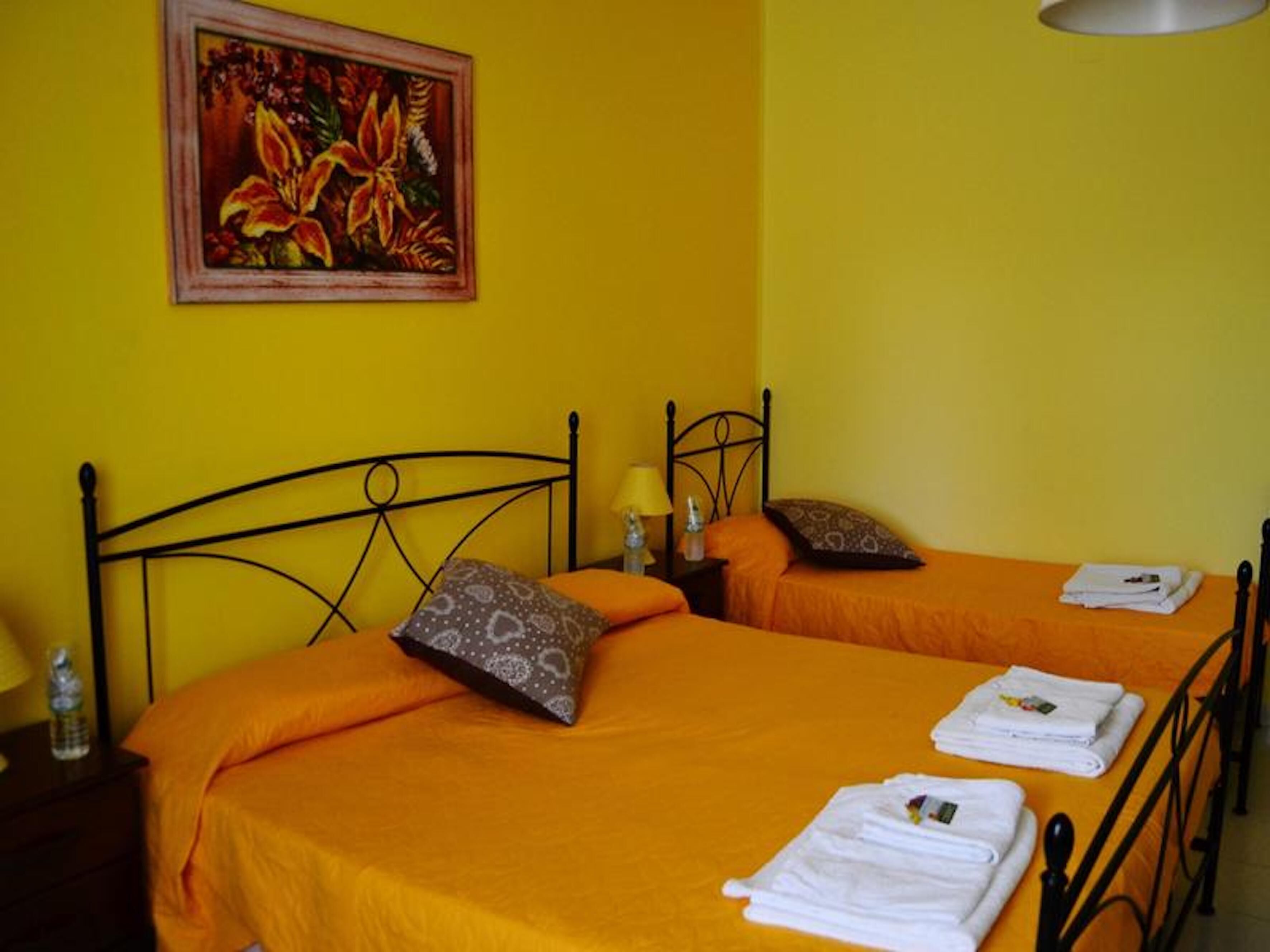 Ein Zimmer mit zwei Betten, jeweils mit orangefarbener Bettdecke und Kissen, einem Bild an der Wand und einer Lampe auf dem Nachttisch.