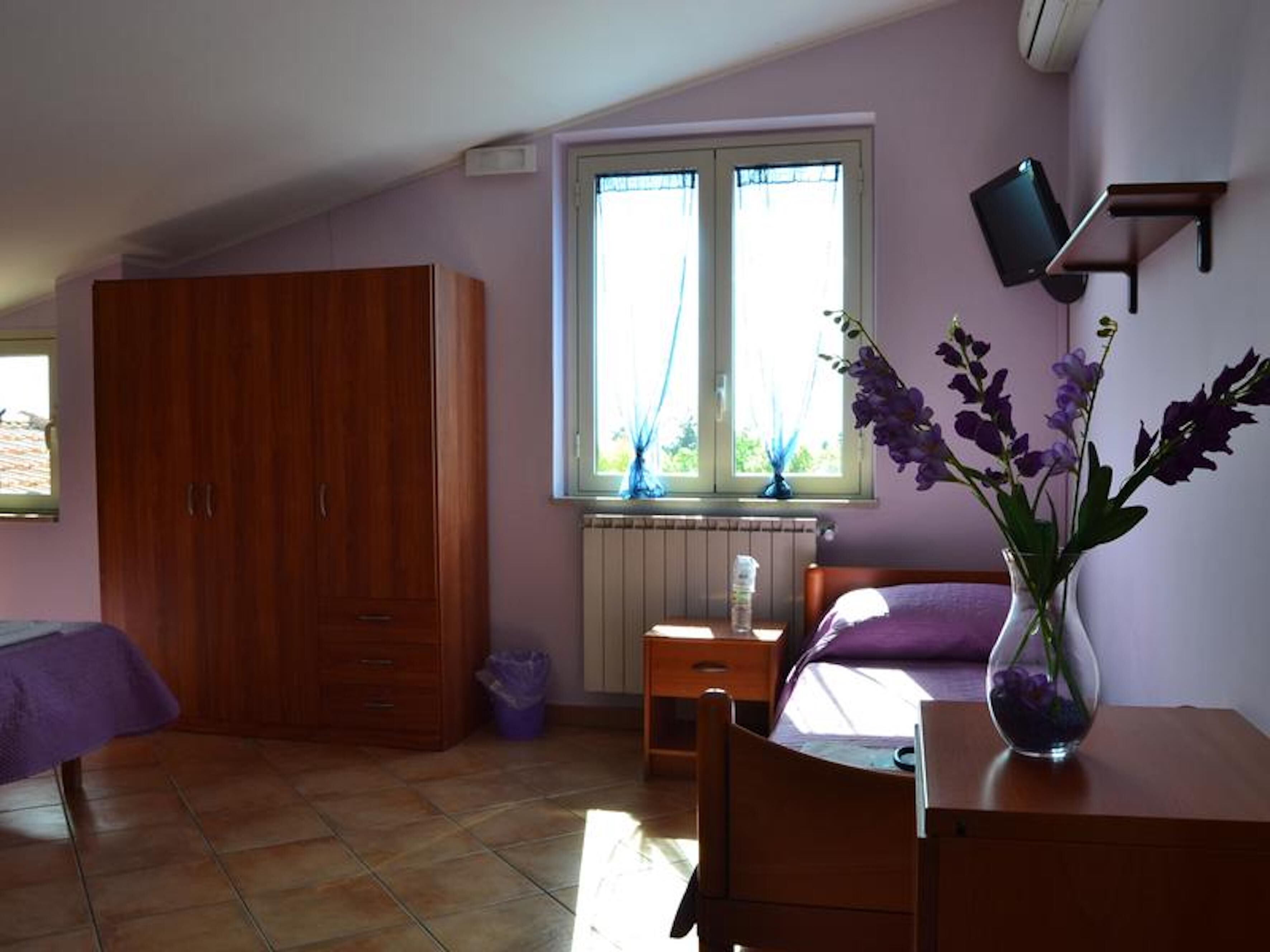 A bedroom with a wooden wardrobe, a bed with a purple bedspread, a small bedside table, a radiator, and a window with curtains.
