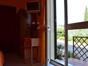 A room with a desk, a chair, a TV mounted on the wall, and a view of a garden through the door. - Room-suite Rosa - B&b S - Elia (Caltanissetta)