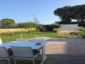 An outdoor patio area with a table and chairs, a paved walkway, and a hedge in the background.