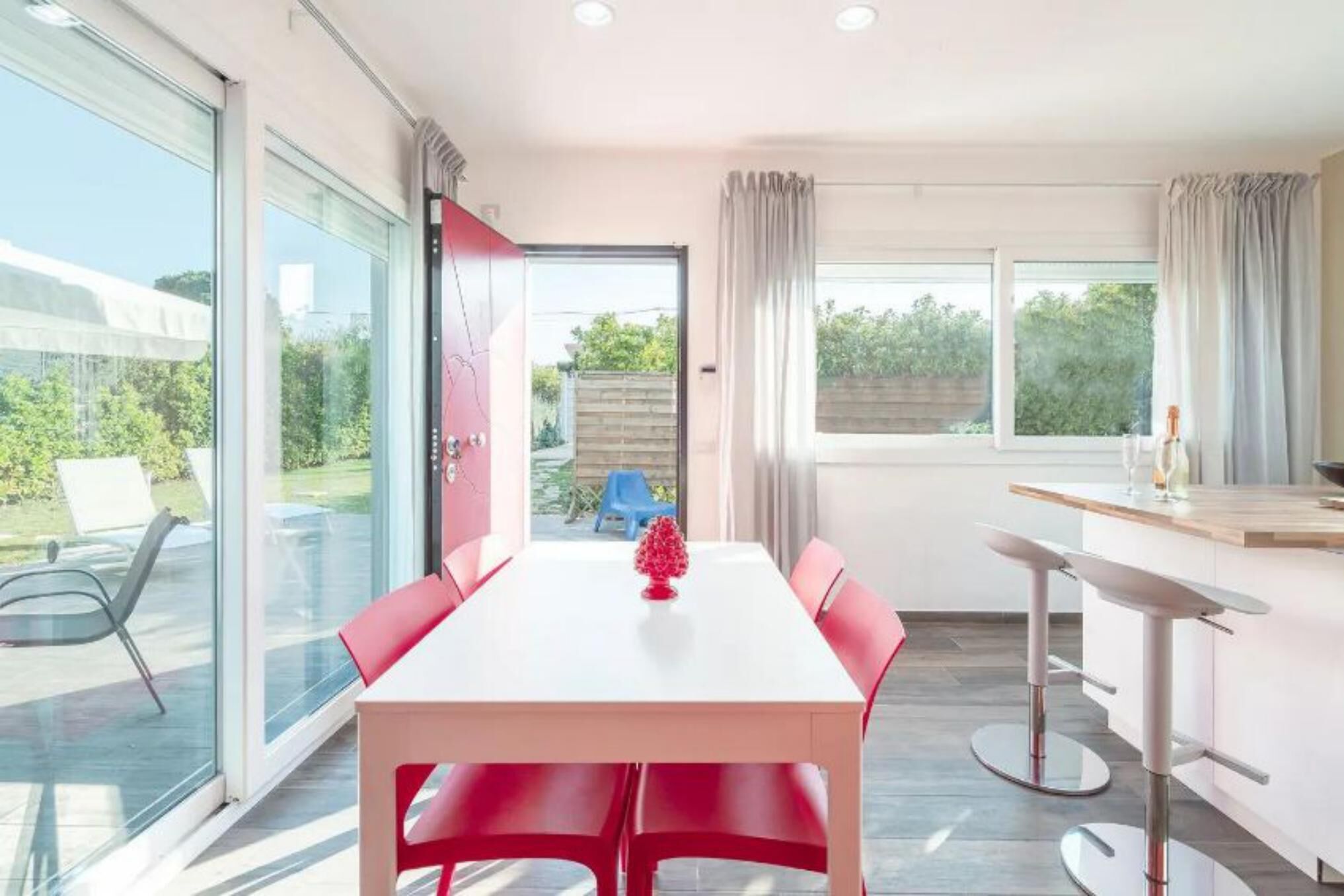 A modern dining area with a white table and red chairs, a glass door leading to a patio, and a kitchen area with a bar counter.