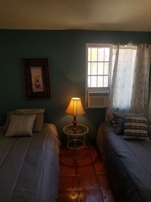 A small, cozy room with two single beds, a table with a lamp, a window with curtains, and a framed picture on the wall. - Beautiful House For Vacation In Ensenada (Ensenada)