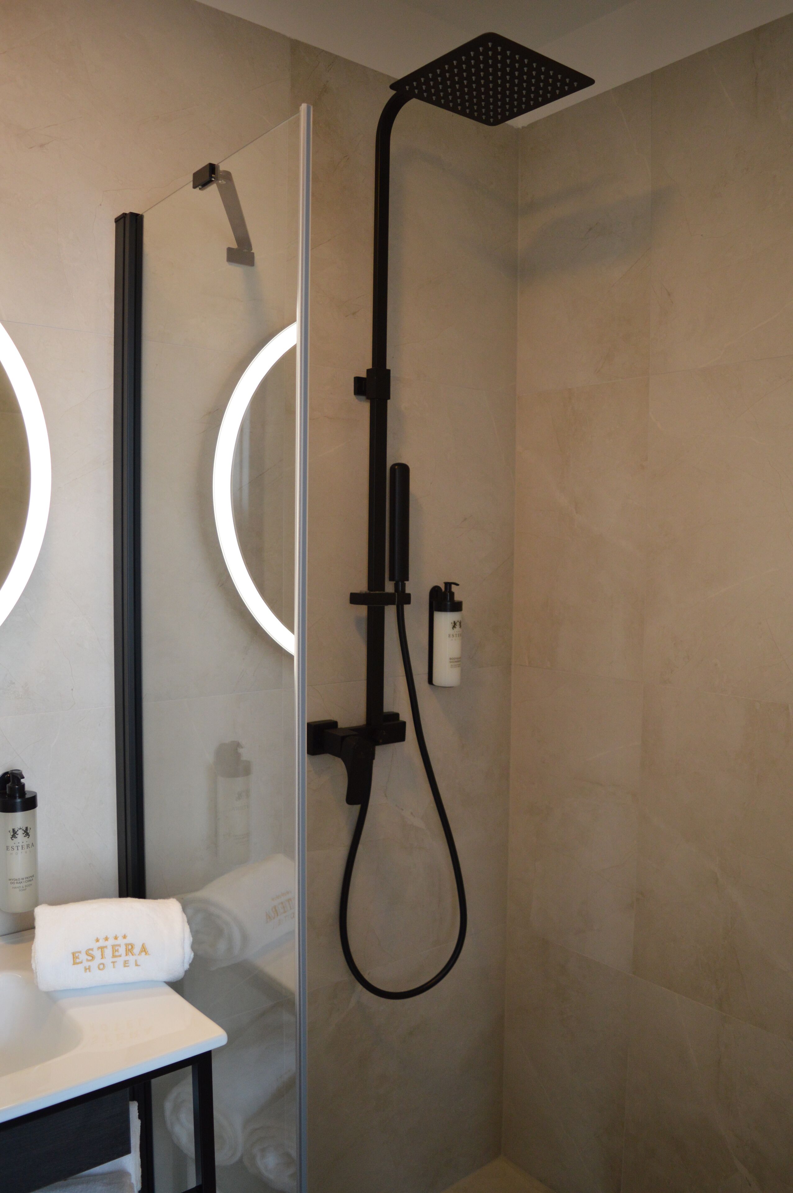 Une salle de bain moderne avec une paroi en verre, une douchette à main et un porte-serviettes sur lequel est posée une serviette portant l’inscription « HÔTEL ESTERA ».