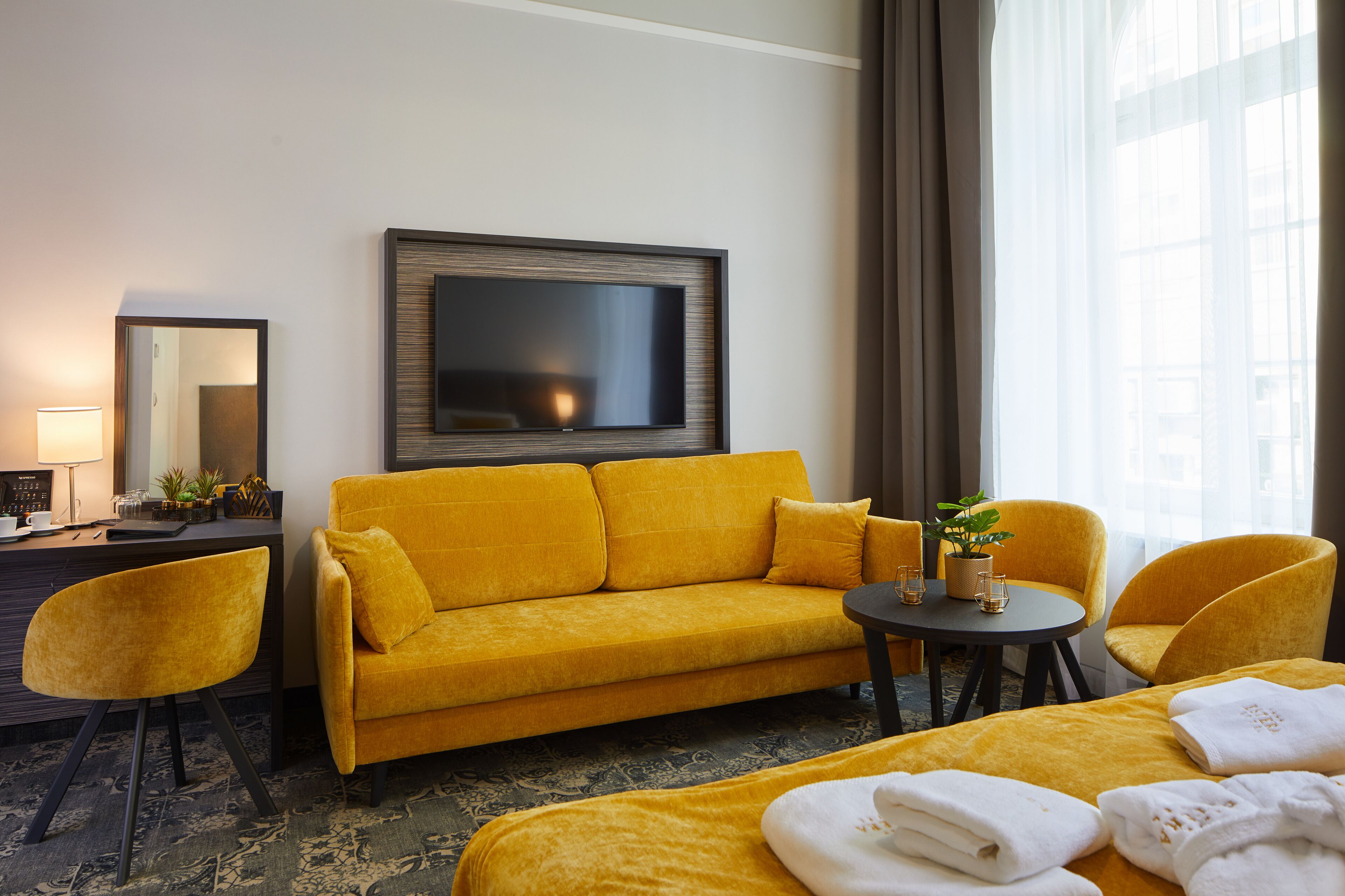 Habitación de hotel con sofá, dos sillones, una mesita con una planta, un televisor de pantalla plana en la pared y una cama con toallas.