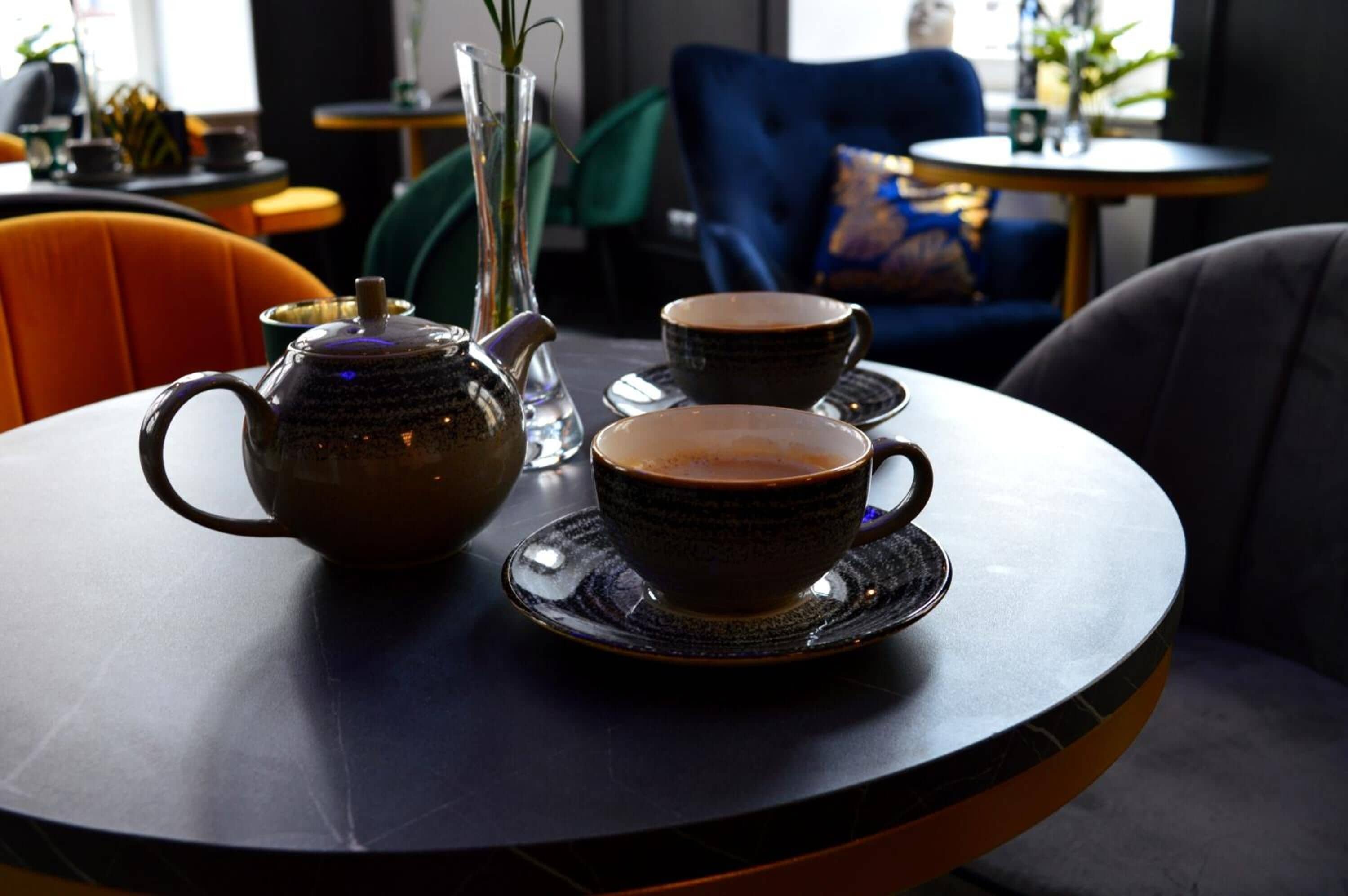 A table with a teapot and two cups, a vase with a plant, and a blue chair in a cafe setting.