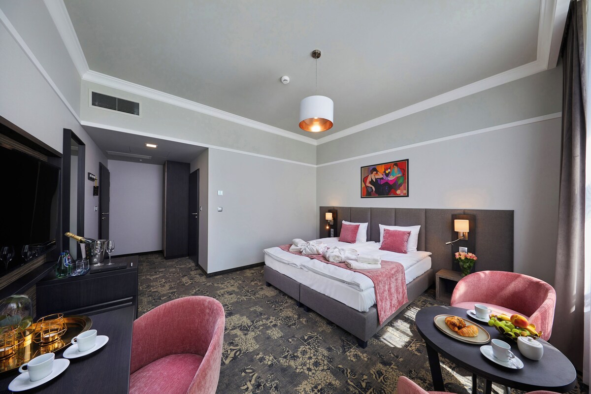 A hotel room with a bed, a desk with a computer, a chair, a table with food and drinks, and a painting on the wall.