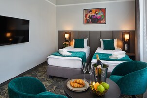 A hotel room with two beds, a flat-screen TV, a painting, a minibar, and a breakfast table with croissants and fruit.