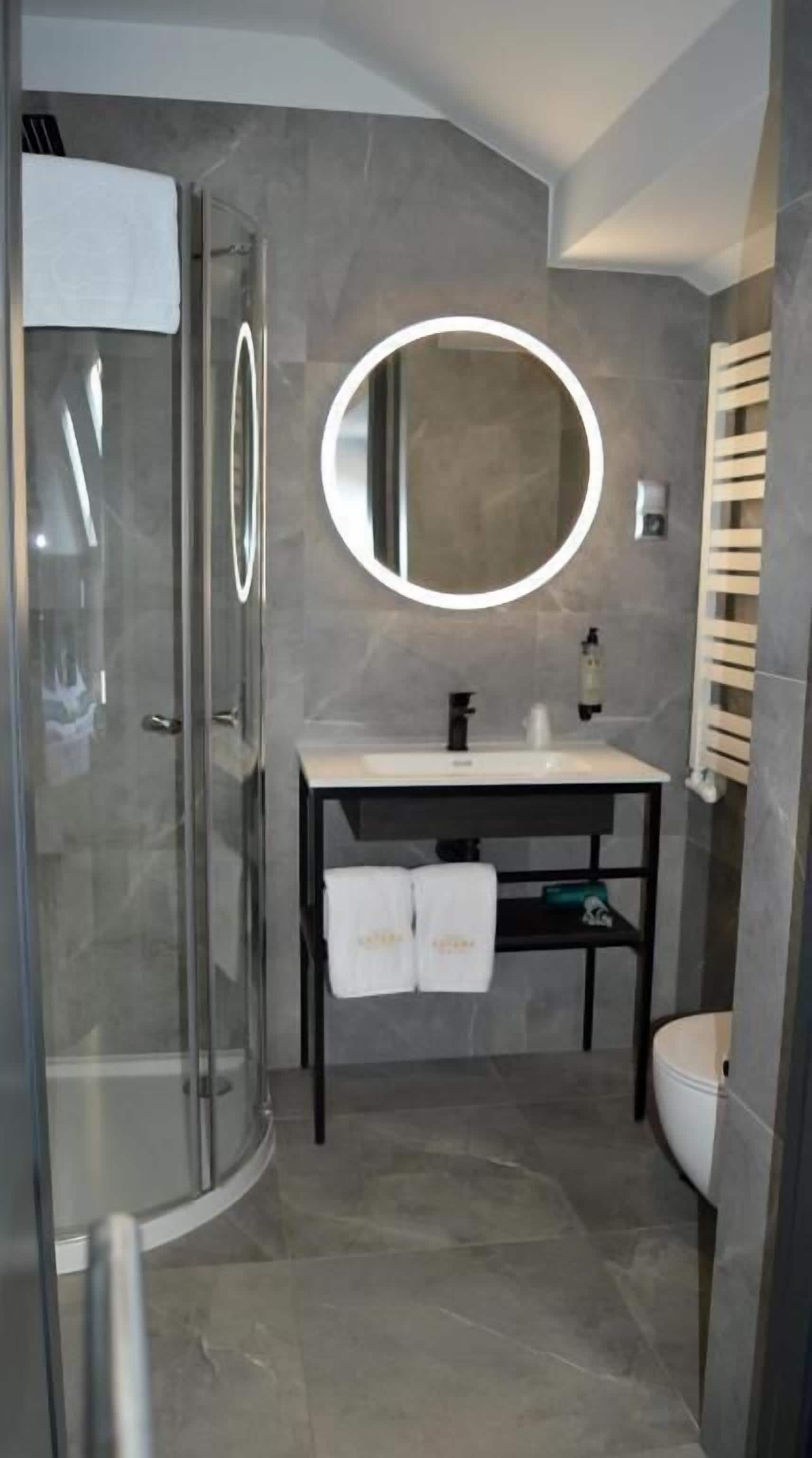 A modern bathroom with a glass shower enclosure, a round mirror, a white sink, and a toilet.