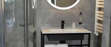 A modern bathroom with a glass shower enclosure, a round mirror, a white sink, and a toilet.