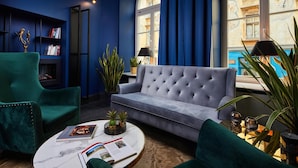 A modern living room with a grey sofa, teal armchairs, and a marble coffee table. There is a bookshelf with books and decorative items, and a large window with curtains. - Hotel Estera (Kraków)