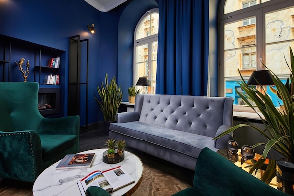 A modern living room with a grey sofa, teal armchairs, and a marble coffee table. There is a bookshelf with books and decorative items, and a large window with curtains.