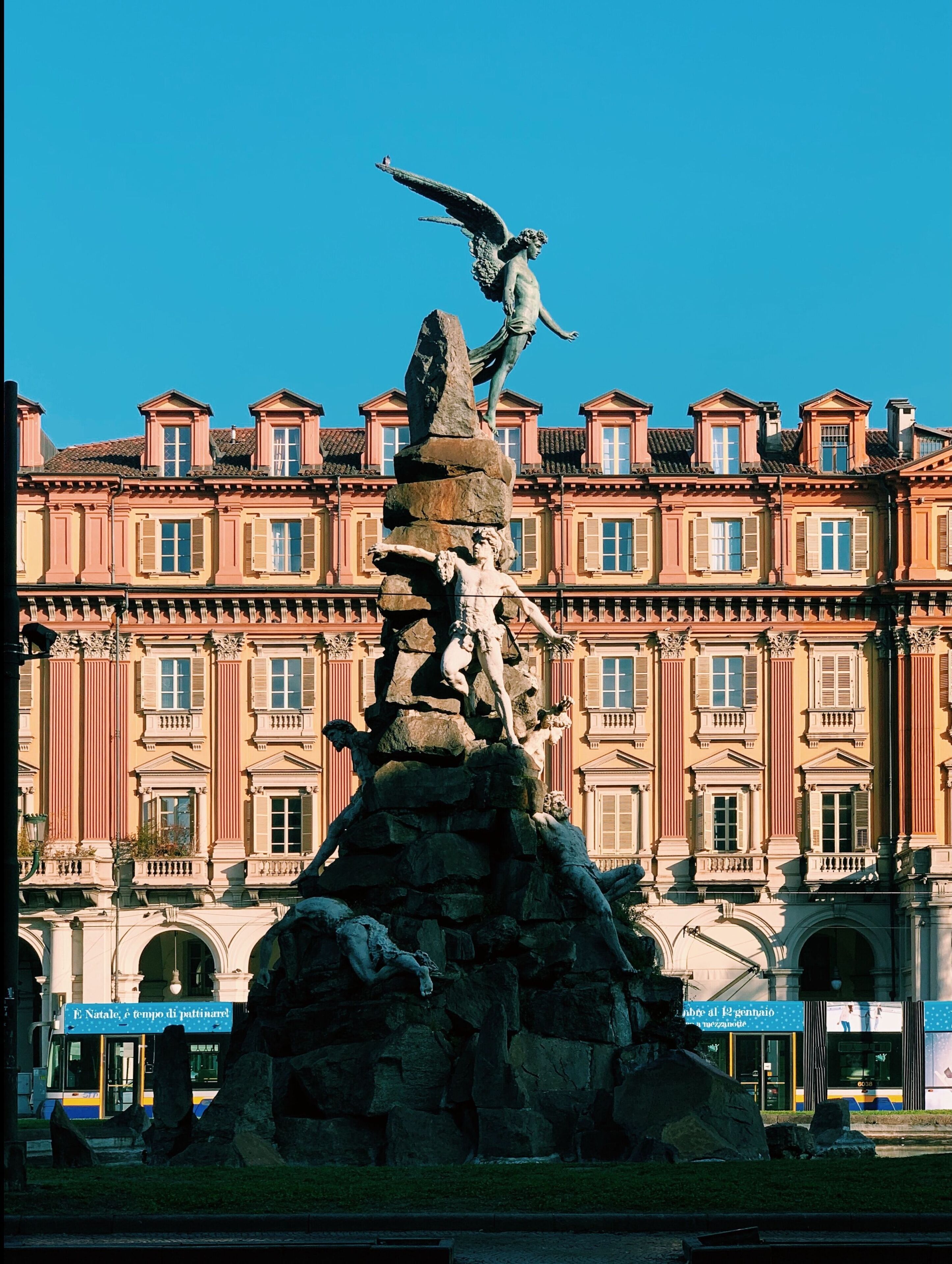 A classical statue atop a rocky pedestal in front of a historic building with multiple windows and balconies.