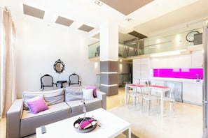 Ein modernes Wohnzimmer mit einer Couch, Sesseln, einem Esstisch und einer Küche mit einer leuchtend rosa Spritzschutzwand.