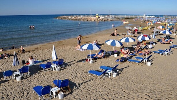 A beach with sun loungers and umbrellas, a pier in the background, and calm water. - Gouves Beach Boutique Apartment (Hersonissos)