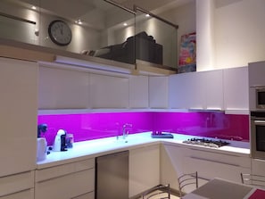 Ett modernt kök med vita skåp, en lila väggbeklädnad bakom arbetsbänken, en inbyggd ugn och en klocka på väggen.