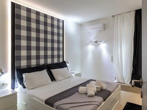 Hotellihuone, jossa on ruutukuviollinen sängynpääty, valkoisilla lakanoiden peitetty sänky, yöpöytä, seinälle asennettu ilmastointilaite ja suihkutila, jossa on verhot.
