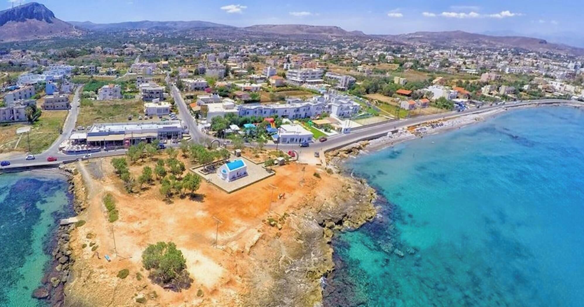 An aerial view of a coastal town with residential buildings, a main road, and a clear blue sea.