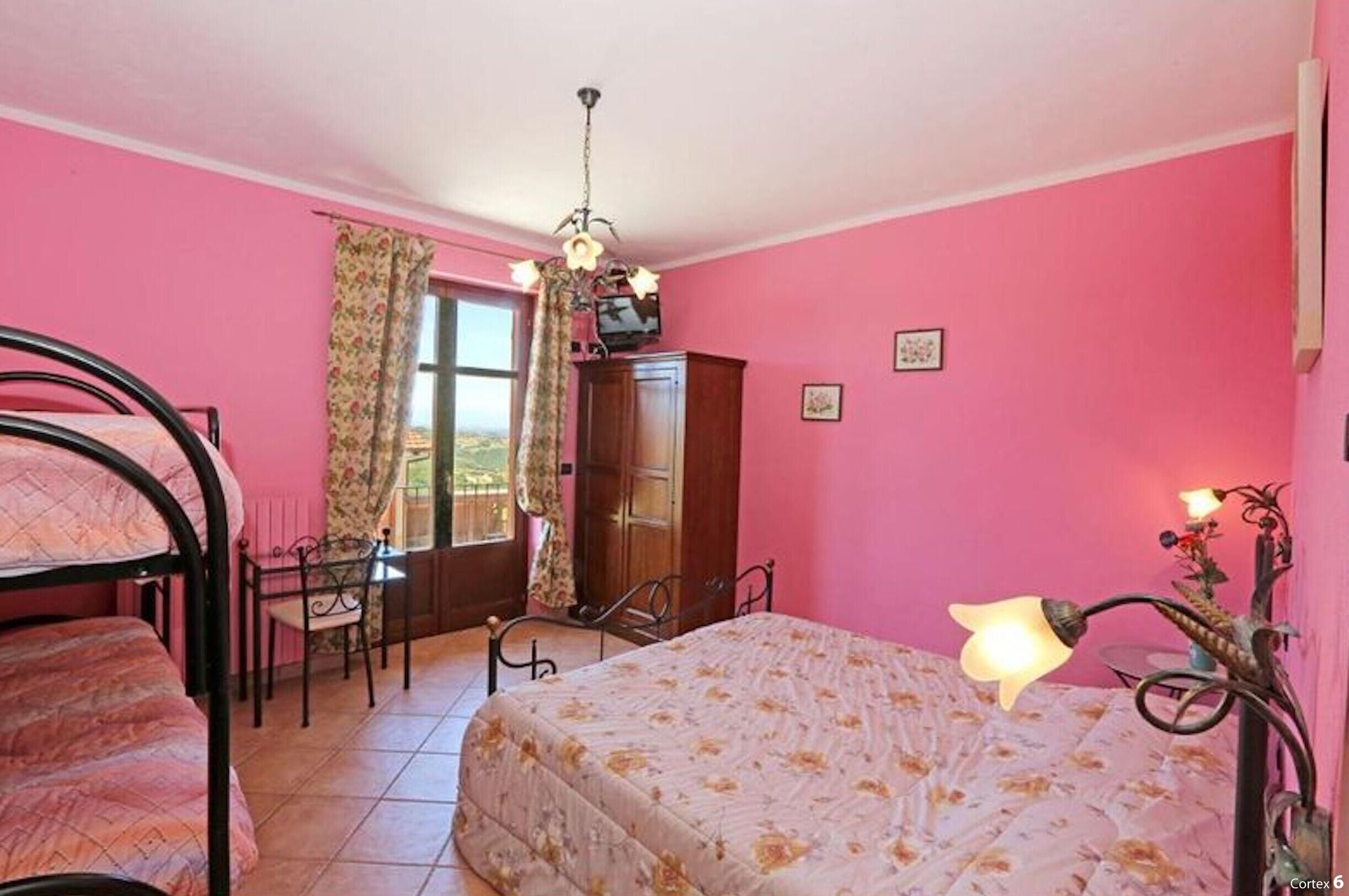 A bedroom with pink walls, a bed with floral bedding, a desk with a chair, a wardrobe, and a window with curtains.