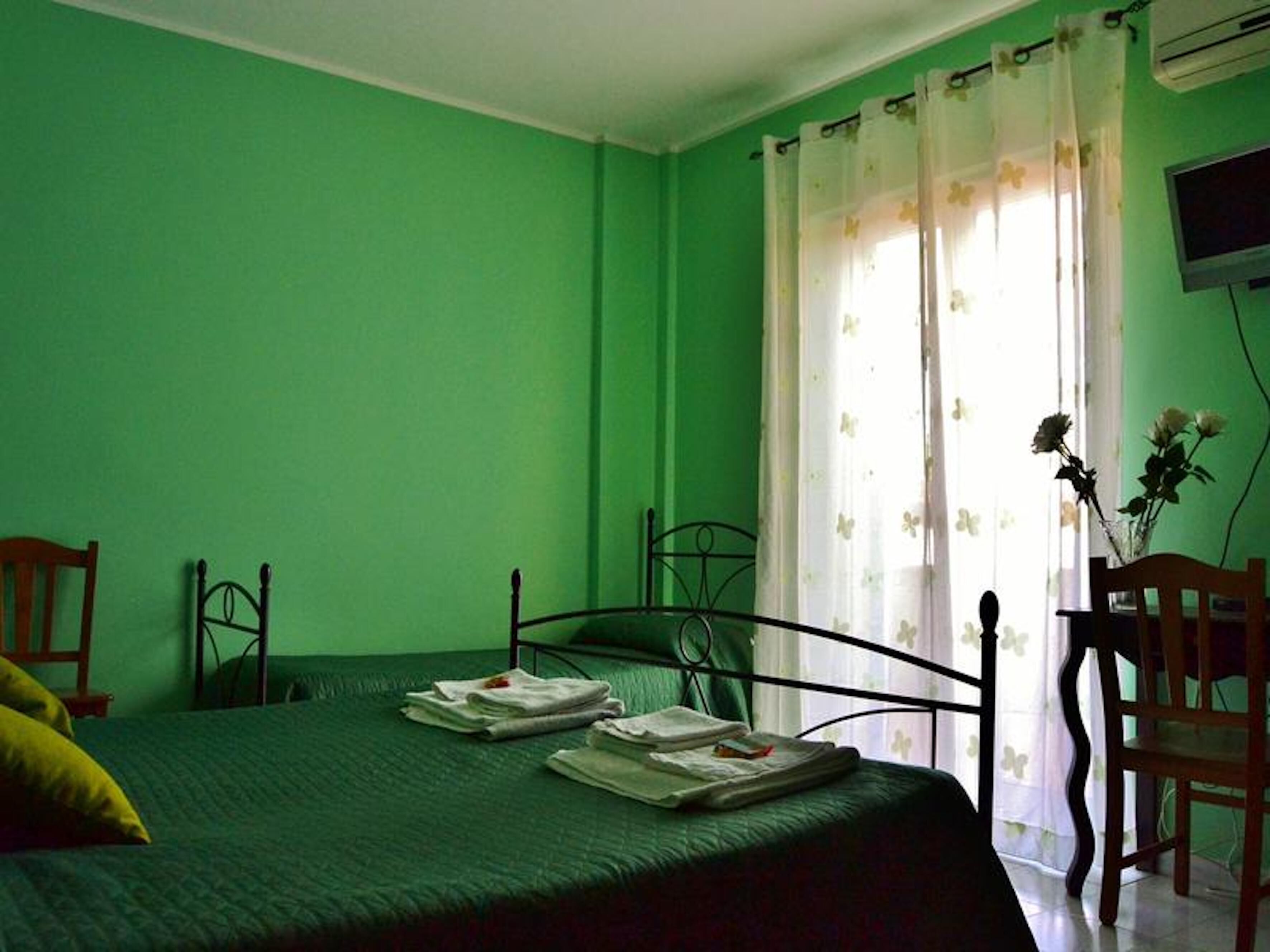 Una habitación con paredes verdes, una cama con edredón verde, dos sillas, un ventanal con cortinas florales y un televisor en la pared.