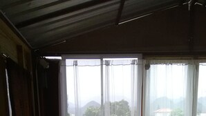 Ein Zimmer mit Fliesenboden, Holzbalkendecke und Blick auf ein Gebäude durch transparente Vorhänge. Plastikstühle und ein Tisch sind zu sehen.