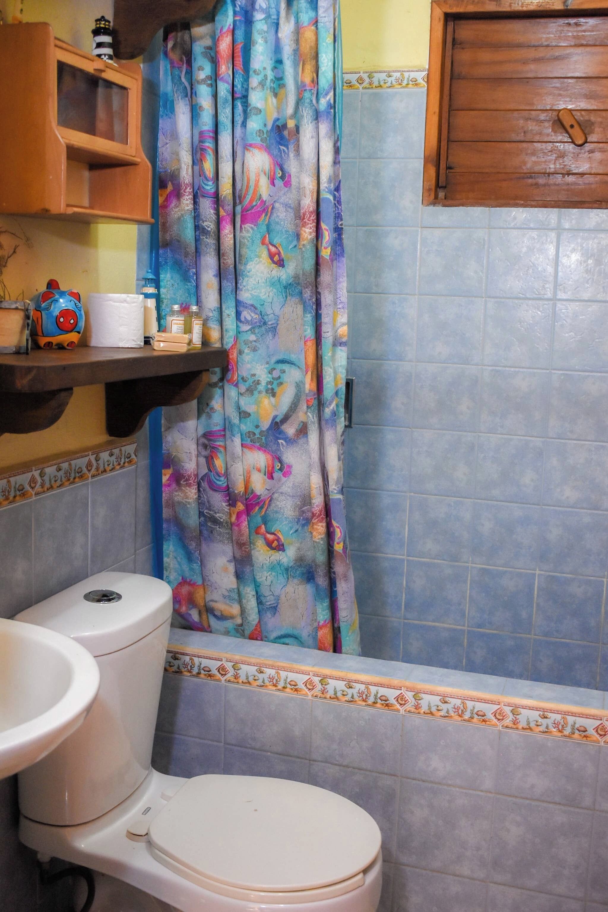 Un baño con inodoro, lavamanos, cortina de ducha con estampado de peces y un armario de madera sobre el lavamanos.