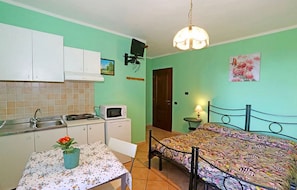 A compact room with a bed, a small table with a vase, a kitchenette with cabinets, a microwave, and a hanging light fixture.