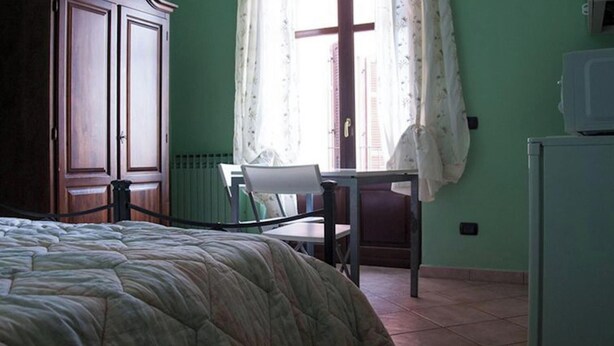 A bedroom with a bed, a chair, a desk, a radiator, and a window with curtains.