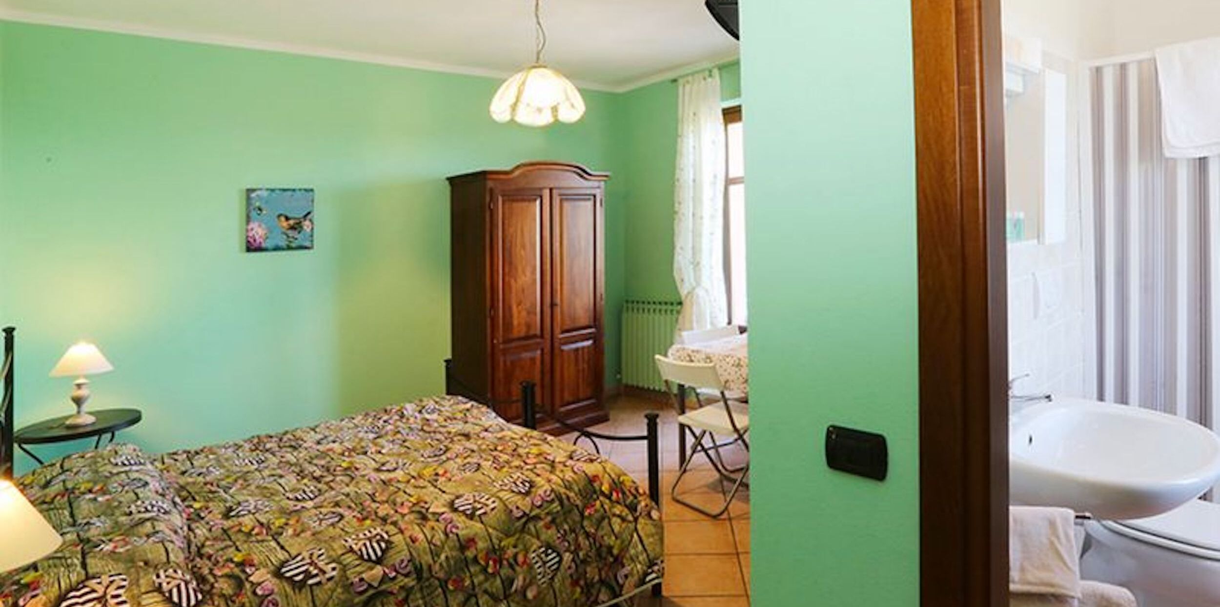 A room with a bed, a wooden cabinet, a table with a lamp, and a bathroom with a sink and toilet.