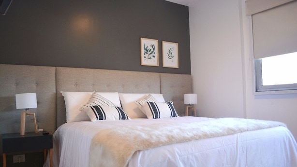 Een net opgemaakt bed met witte lakens en gestreepte kussens, met nachtkastjes met lampen ernaast, in een kamer met een grijze hoofdborden en twee ingelijste botanische prints aan de muur.