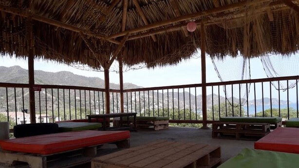 Un espacio al aire libre con bancos y mesas de madera, techo de paja y vistas a las montañas y al mar.