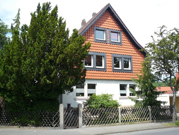 A two-story house with a red-tiled roof and white walls, surrounded by a wooden fence and trees.