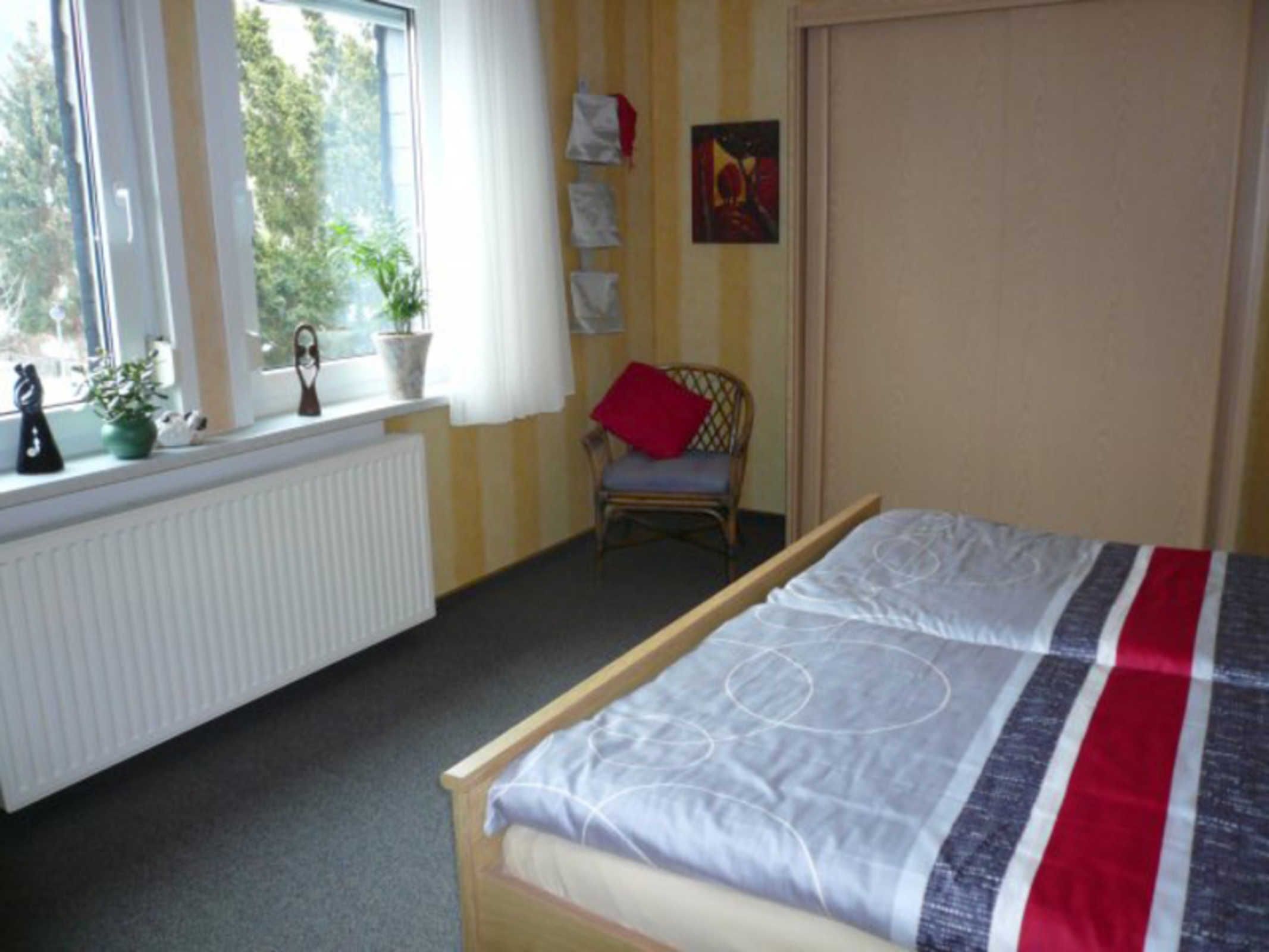 Une chambre à coucher comprenant un lit, une chaise, un radiateur, une fenêtre avec des rideaux et un mur orné de tableaux.