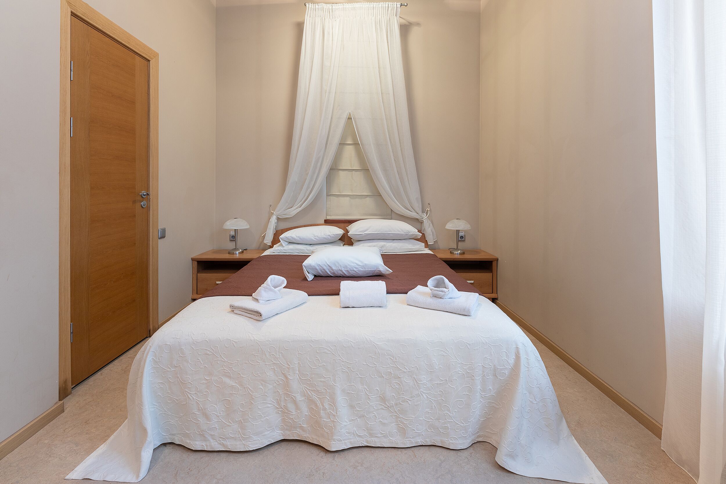Una cama bien tendida con sábanas y toallas blancas, una puerta de madera y una ventana con cortina translúcida.