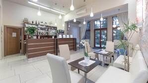 A modern reception area with a wooden desk, white chairs, and a reception counter. There are hanging lights and a large window providing natural light. - Room in Guest Room - Valensija - Standard Twin Room (Jurmala)