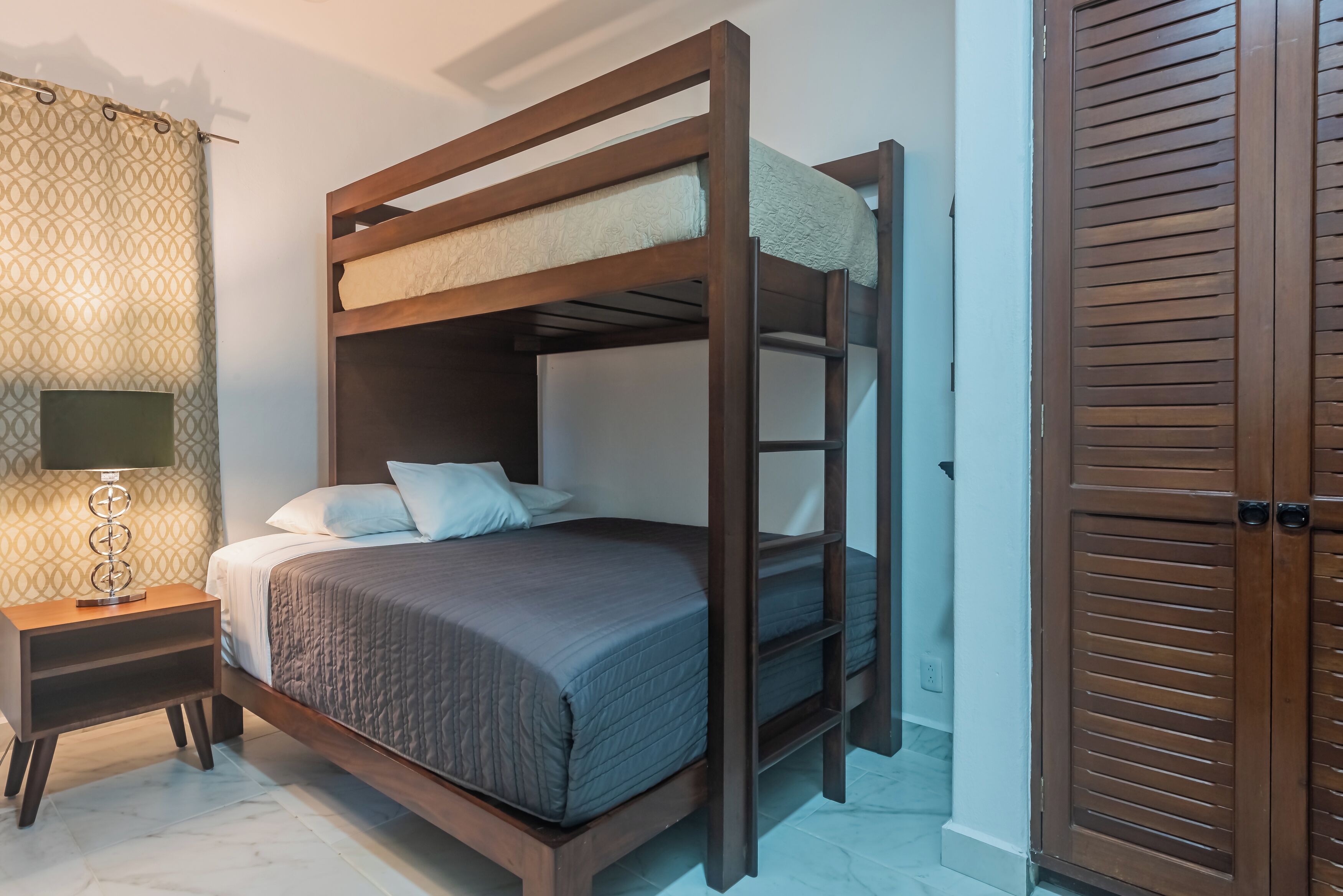 A bunk bed with a wooden frame and a headboard, a bedside table with a lamp, and a patterned curtain.