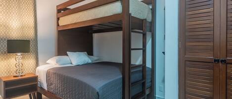 A bunk bed with a wooden frame and a headboard, a bedside table with a lamp, and a patterned curtain.