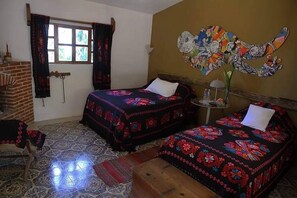 A room with two beds, a brick fireplace, a window with curtains, and a colorful wall decoration. - Ideal Para Familia Pequena O Grupo De Tres Amigos (Cuetzalan del Progreso)