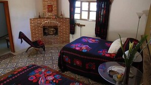 A room with a brick fireplace, two beds with colorful bedding, a patterned rug, and a small table with a vase. - Ideal Para Familia Pequena O Grupo De Tres Amigos (Cuetzalan del Progreso)