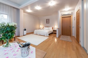 A hotel room with a bed, bedside tables, a nightstand, a large window with curtains, a potted plant, and a minibar. - Room in Guest Room - Valensija - Large Suite Apartment (Jurmala)