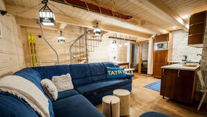A cozy room with a blue sofa, wooden flooring, a kitchen area, and ski equipment.