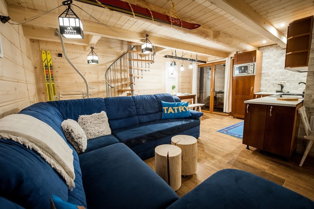 A cozy room with a blue sofa, wooden flooring, a kitchen area, and ski equipment.