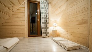 A room with wooden walls and a sloped ceiling, a wooden door, a patterned curtain, a small bedside table with a lamp, and two pet beds.