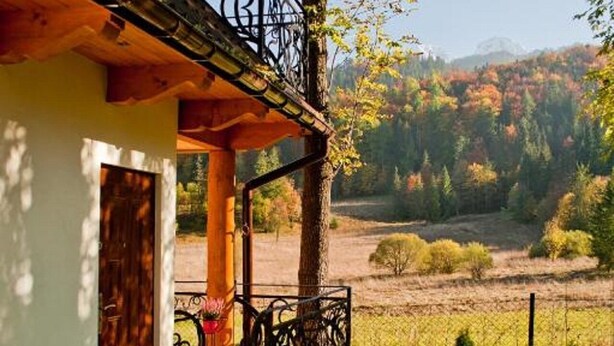 A rustic house with a balcony, a wooden door, and a small garden area with a fence and trees.