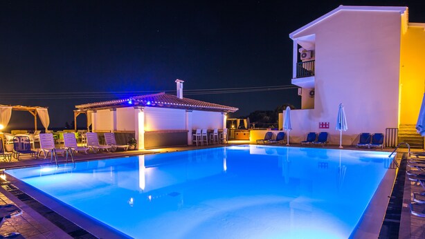 Una piscina con sillas de descanso y sombrillas, rodeada de edificios con balcones y una zona cubierta con luces.