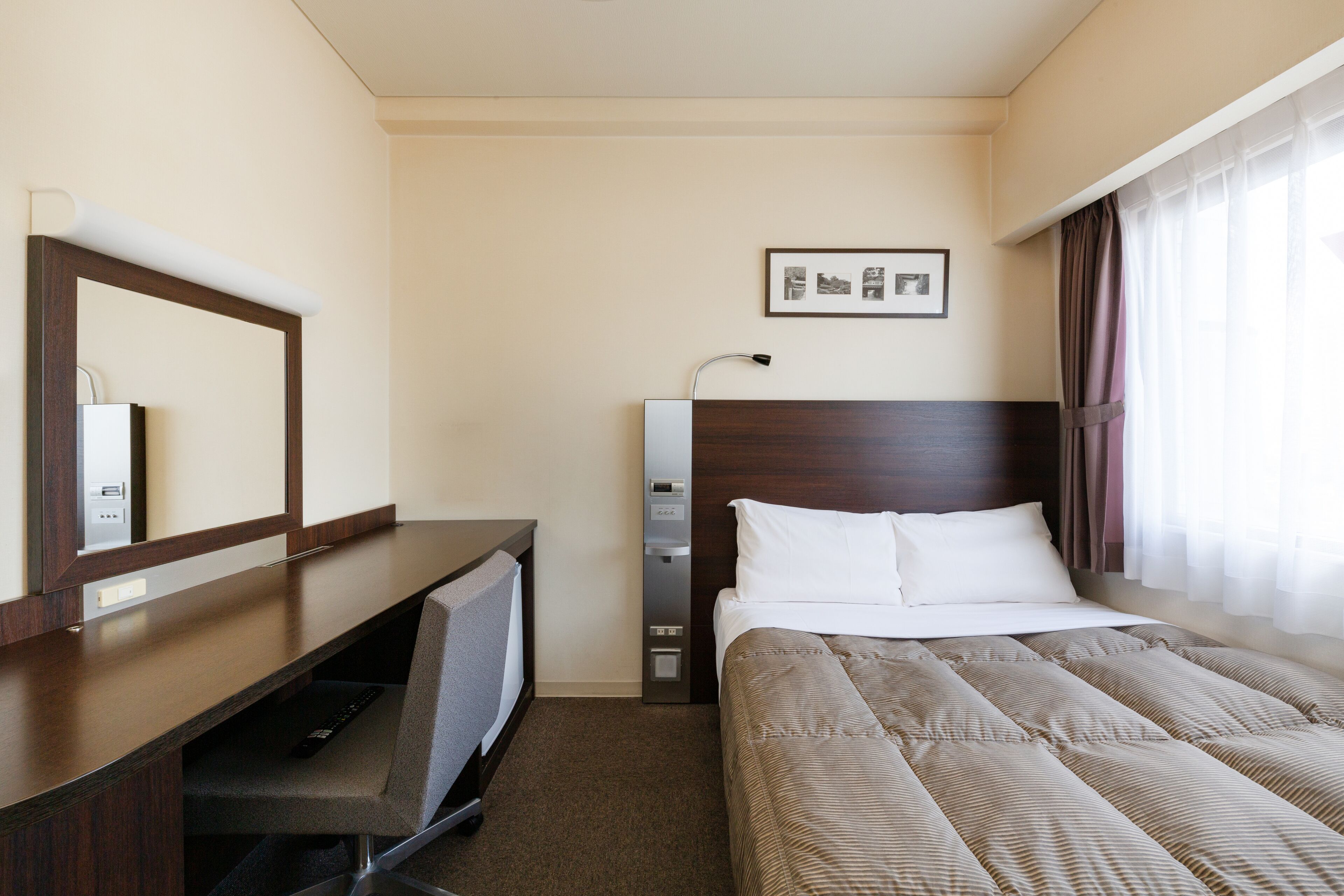 A hotel room with a bed, a desk, a chair, a mirror, a picture on the wall, and a window with curtains.